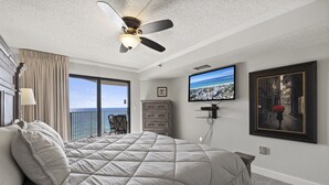 A bedroom with a large bed, a ceiling fan, a TV, and a view of the ocean.