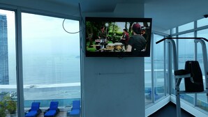Une chambre moderne avec un grand téléviseur fixé au mur, un balcon avec des chaises bleues et une vue sur la mer et les bâtiments.