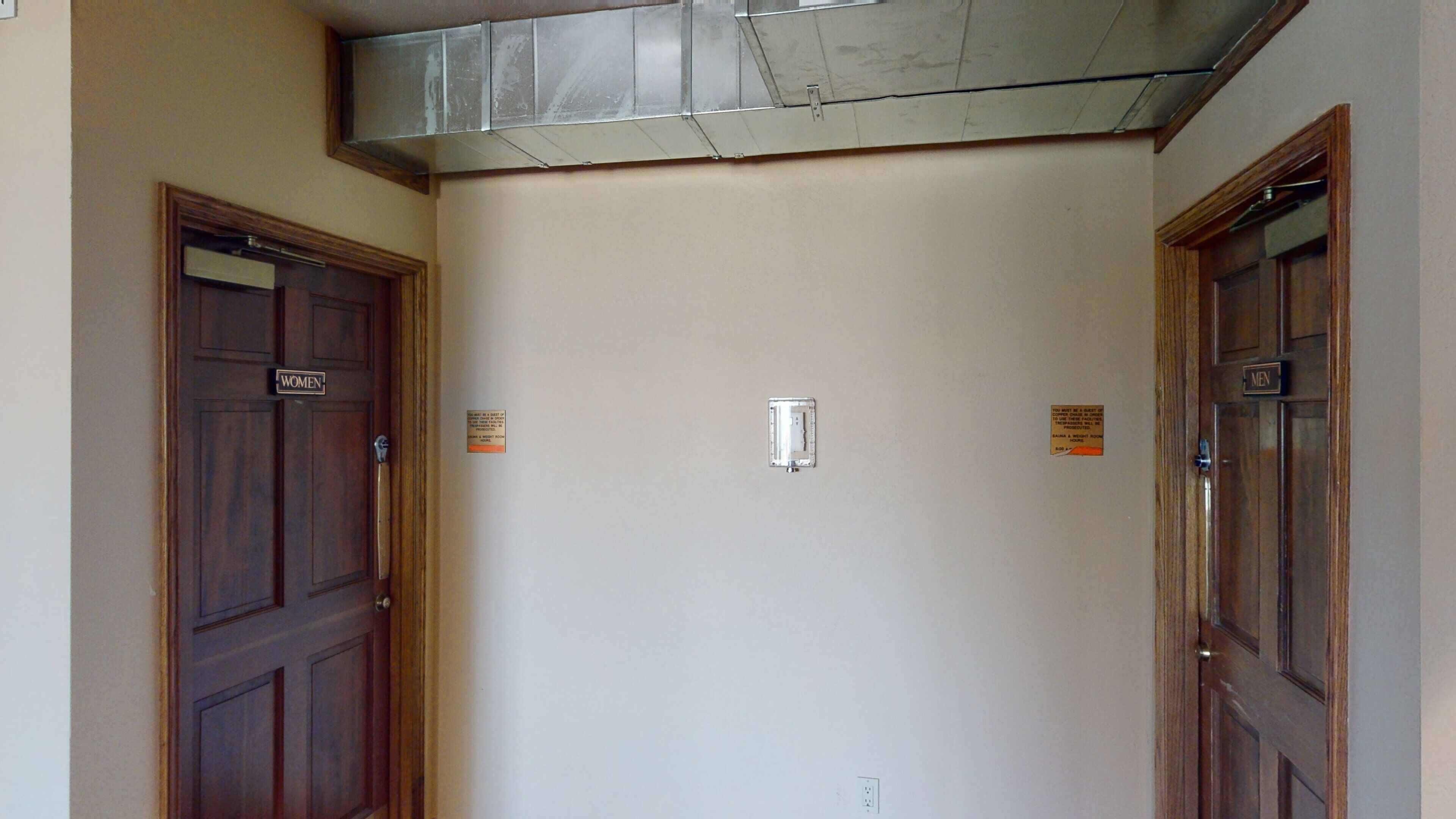 A hallway with two wooden doors labeled "WOMEN" and "MEN" and a small electrical outlet on the wall.