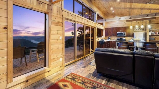 Una cabaña de madera con un gran ventanal corredera que da a una terraza con vistas a las montañas. En el interior, hay una cocina con armarios de madera y un comedor con un sofá.