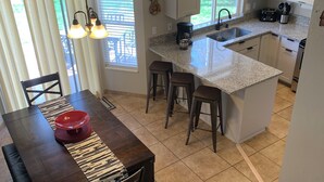 A kitchen with a central island, bar stools, a dining table with chairs, a coffee maker, and a view of the outdoors.