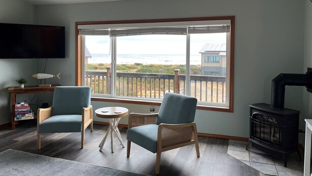 A living room with two blue chairs, a small round table, a TV mounted on the wall, and a large window with a view of the beach and a house.