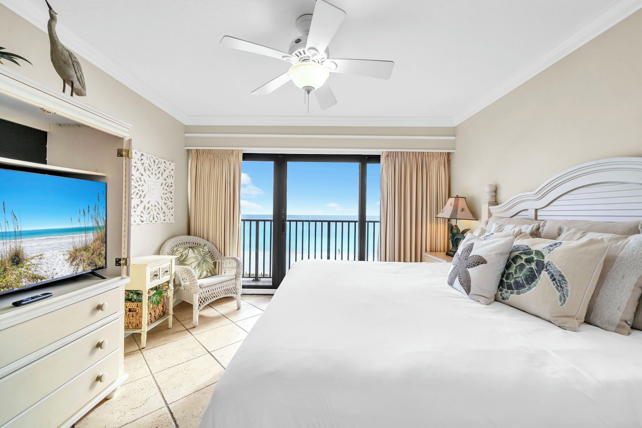 A hotel room with a bed, a TV, a ceiling fan, a balcony with a view of the beach, and a decorative wall piece.