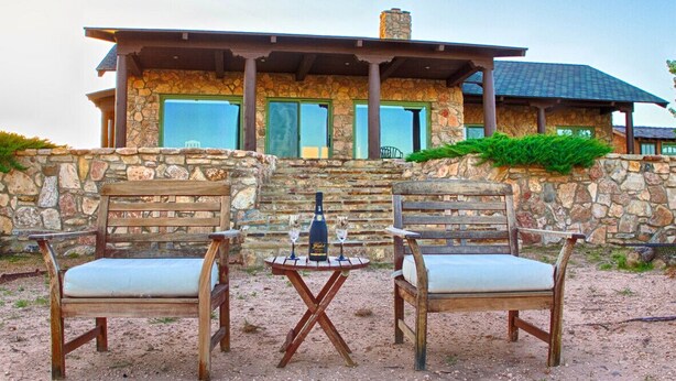 A stone building with a stone wall and a wooden deck. There are two wooden chairs and a small table with a bottle and glasses on it. - SkyHouse cabin on the West Rim (Canyon)