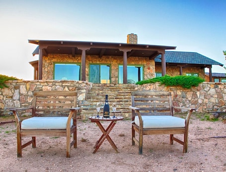 A stone building with a stone wall and a wooden deck. There are two wooden chairs and a small table with a bottle and glasses on it.