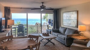A room with a balcony overlooking the ocean, furnished with a sofa, chairs, a table, and a ceiling fan.