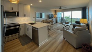 A modern kitchen with white cabinets, a central island, and stainless steel appliances. There is a dining area with a table and chairs, and a living room with a sofa and a TV.