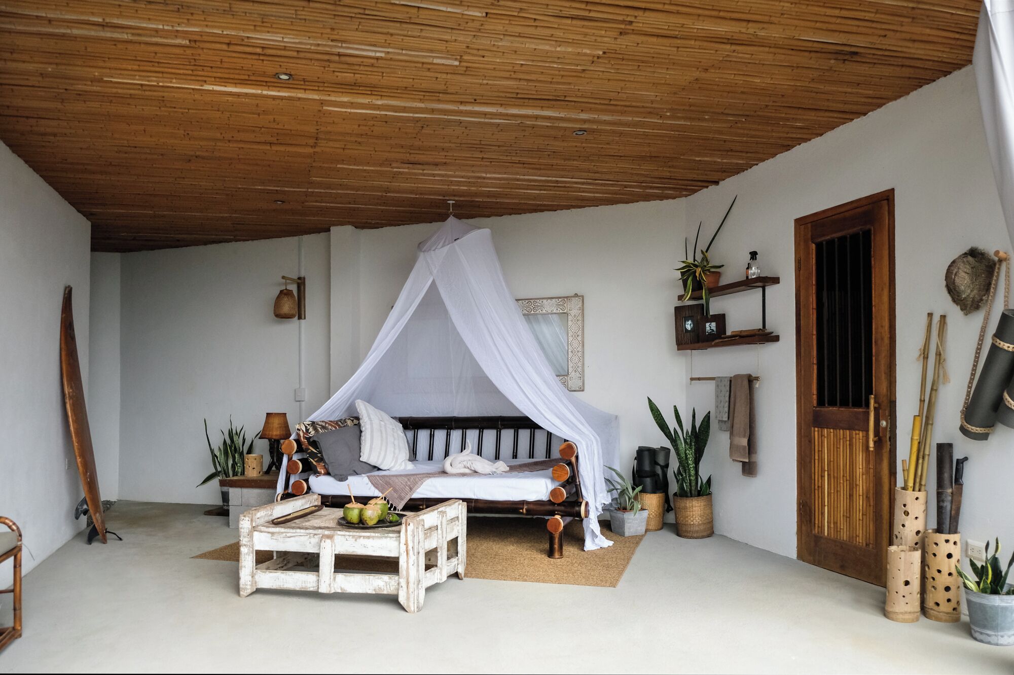 A room with a bed under a canopy, a wooden ceiling, a small table with a plant, and a door with a bamboo door frame.