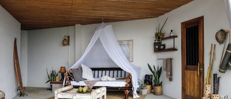 A room with a bed under a canopy, a wooden ceiling, a small table with a plant, and a door with a bamboo door frame.