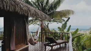 Um deck de madeira com uma rede e uma espreguiçadeira, com vista para uma paisagem exuberante com palmeiras e o horizonte.
