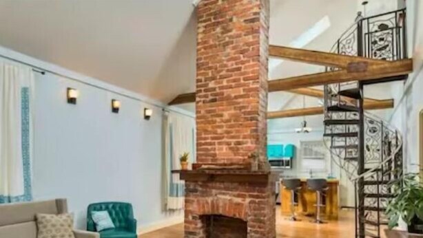 Amplio salón con chimenea de ladrillo, piso de madera, un sofá y una escalera con barandilla de hierro forjado.