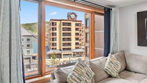 Una sala de estar moderna con un amplio ventanal, un sofá color beige y vistas a un edificio de varios pisos con un reloj.