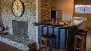 A bar area with a stone fireplace, a clock, and a kitchen in the background.