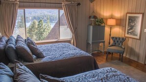 A bedroom with two beds, a view of a forest and lake, a lamp, and a chair.