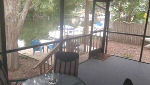 Een overdekt buitengebied met een tafel, stoelen, een houten terras en uitzicht op een waterpartij met boten.