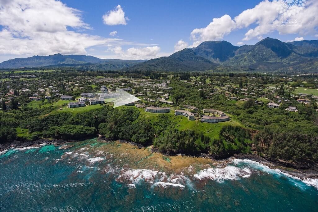 Vista aérea de un complejo turístico costero con varias construcciones, abundante vegetación y montañas al fondo.