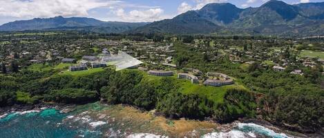 Vista aérea de un complejo turístico costero con varias construcciones, abundante vegetación y montañas al fondo.