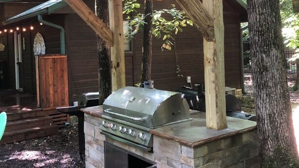Un área de parrilla al aire libre con una barbacoa de piedra, una pérgola de madera y luces colgantes.