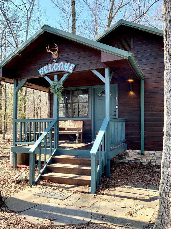 Eine Holzhütte mit einem 'WELCOME'-Schild, einer Veranda mit einer Bank und einer Treppe zum Eingang.