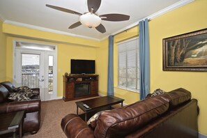 A living room with a ceiling fan, a television on a wooden cabinet, a brown leather sofa, and a painting on the wall. - Luxury Spacious Riverfront Condo in Perfect Location near Tanger Outlets (Sevierville)