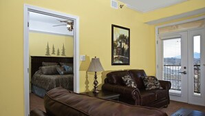 A living room with a brown leather sofa, a small table with a lamp, a bed with a headboard, and a painting on the wall.