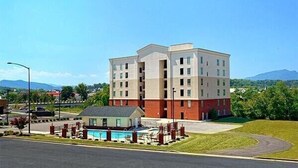 A multi-story hotel with a pool area, located in a suburban setting with a clear sky and mountains in the background.