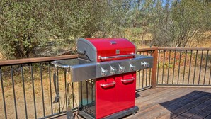A red barbecue grill with four burners and a side shelf, located on a wooden deck with a metal railing. - This quaint cottage is the perfect home base for your Yosemite trip. (Mariposa)
