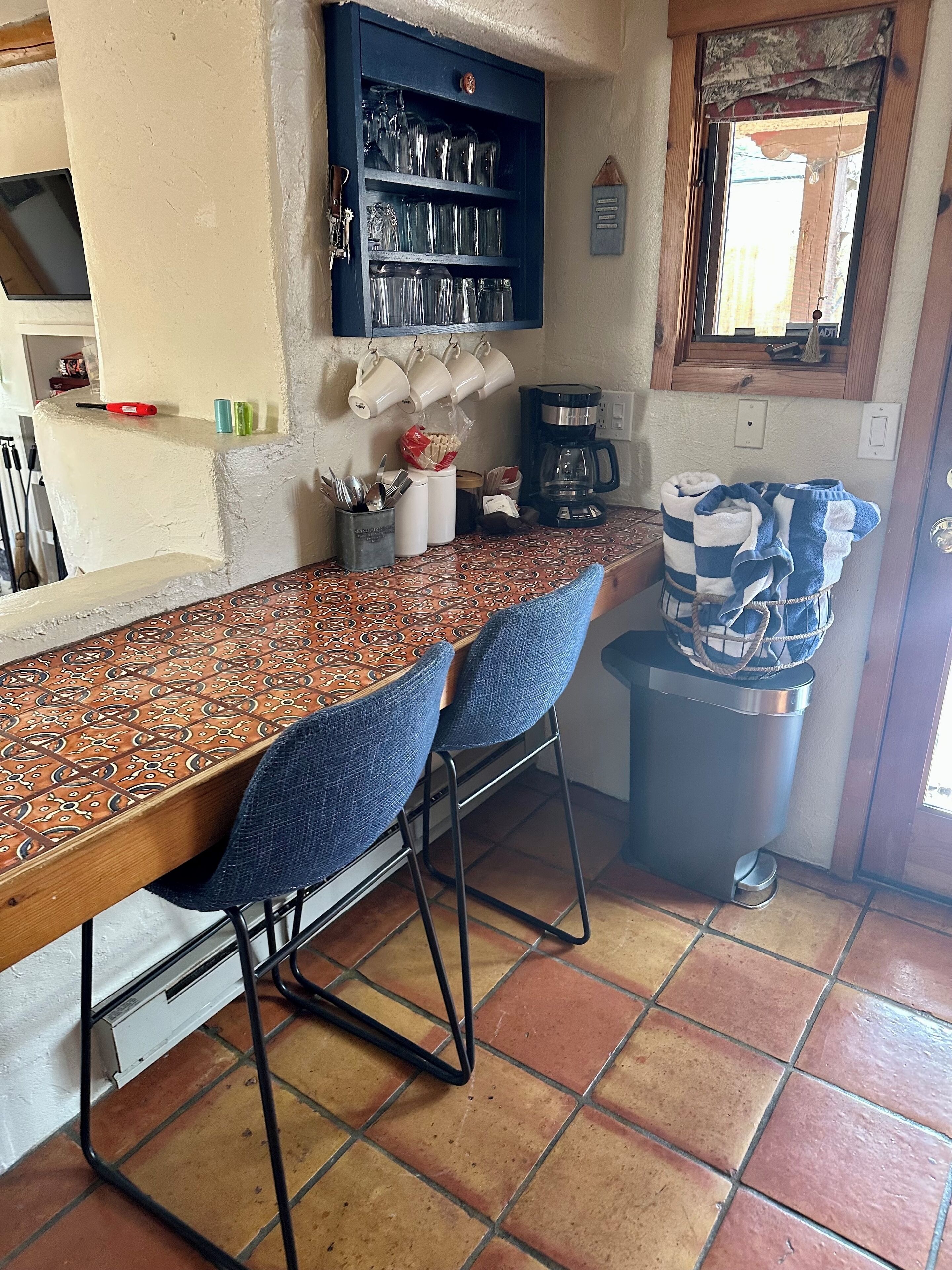 A kitchen area with a long counter, two blue bar stools, a coffee maker, and a trash bin.
