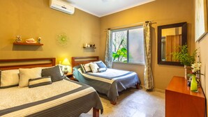 Une chambre d’hôtel avec deux lits, un ventilateur de plafond, un climatiseur, une fenêtre avec des rideaux, un miroir et une petite table avec un vase.