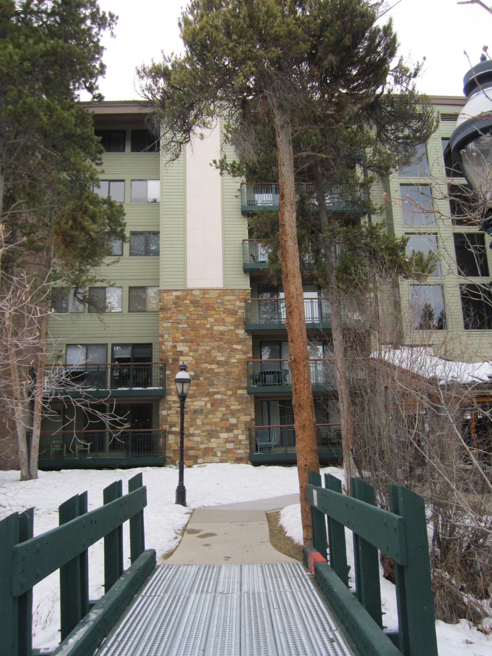 A multi-story building with a stone base and green siding, surrounded by trees and a snow-covered ground.