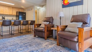 A living room with a wooden coffee table, two leather armchairs, a blue patterned rug, and a kitchen in the background. - Best Location in Town! Ski-in/Ski-out on Peak 9! (Breckenridge)