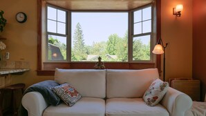 A room with a white sofa, two decorative pillows, a clock on the wall, and a window with a view of trees and a house. - The Ashland Cottage, walk to town (Ashland)