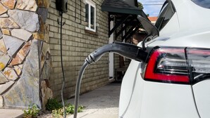 En bil parkeret foran et hus med en stenvæg og en sort el-boks fastgjort til væggen.
