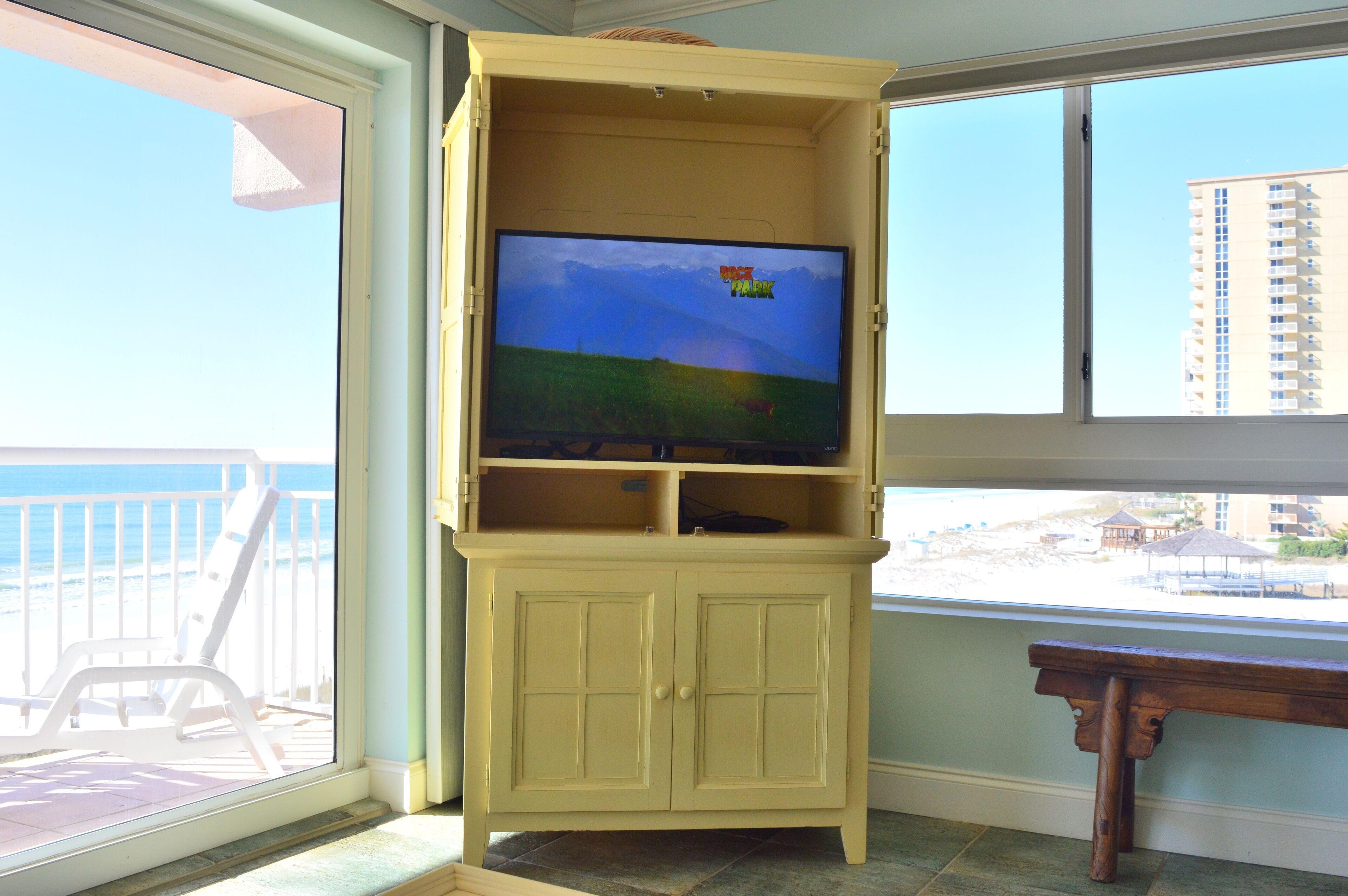 A room with a large TV on a yellow cabinet, a balcony with a chair, and a view of the beach and buildings.
