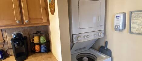 A compact kitchen with wooden cabinets, a coffee maker, a washing machine, and a basket on the counter.