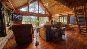 A log cabin interior with a wooden ceiling, a television on the wall, a sofa, a coffee table, and a dining table.
