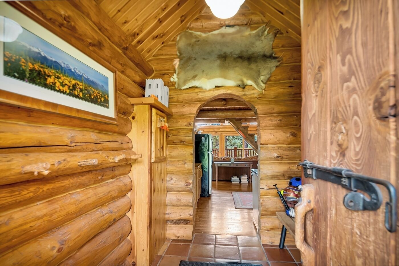 A log cabin interior with a wooden door, a framed picture on the wall, and a view into the living area.