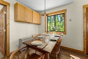 Un coin repas avec une table en bois dressée pour six personnes, des placards en bois au-dessus et une fenêtre donnant sur la forêt.