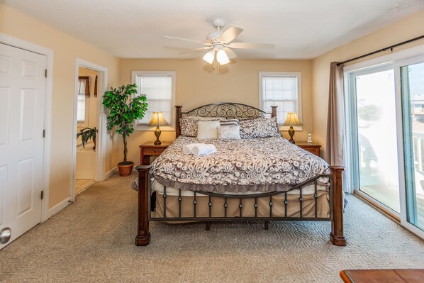 A bedroom with a bed, ceiling fan, two bedside lamps, a potted plant, and a view through a large window.