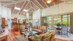 A spacious living area with a high ceiling, wooden flooring, and a large window offering a view of greenery.