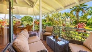 A covered outdoor seating area with wicker furniture and cushions, overlooking a lush garden with tropical plants and a mountain view.