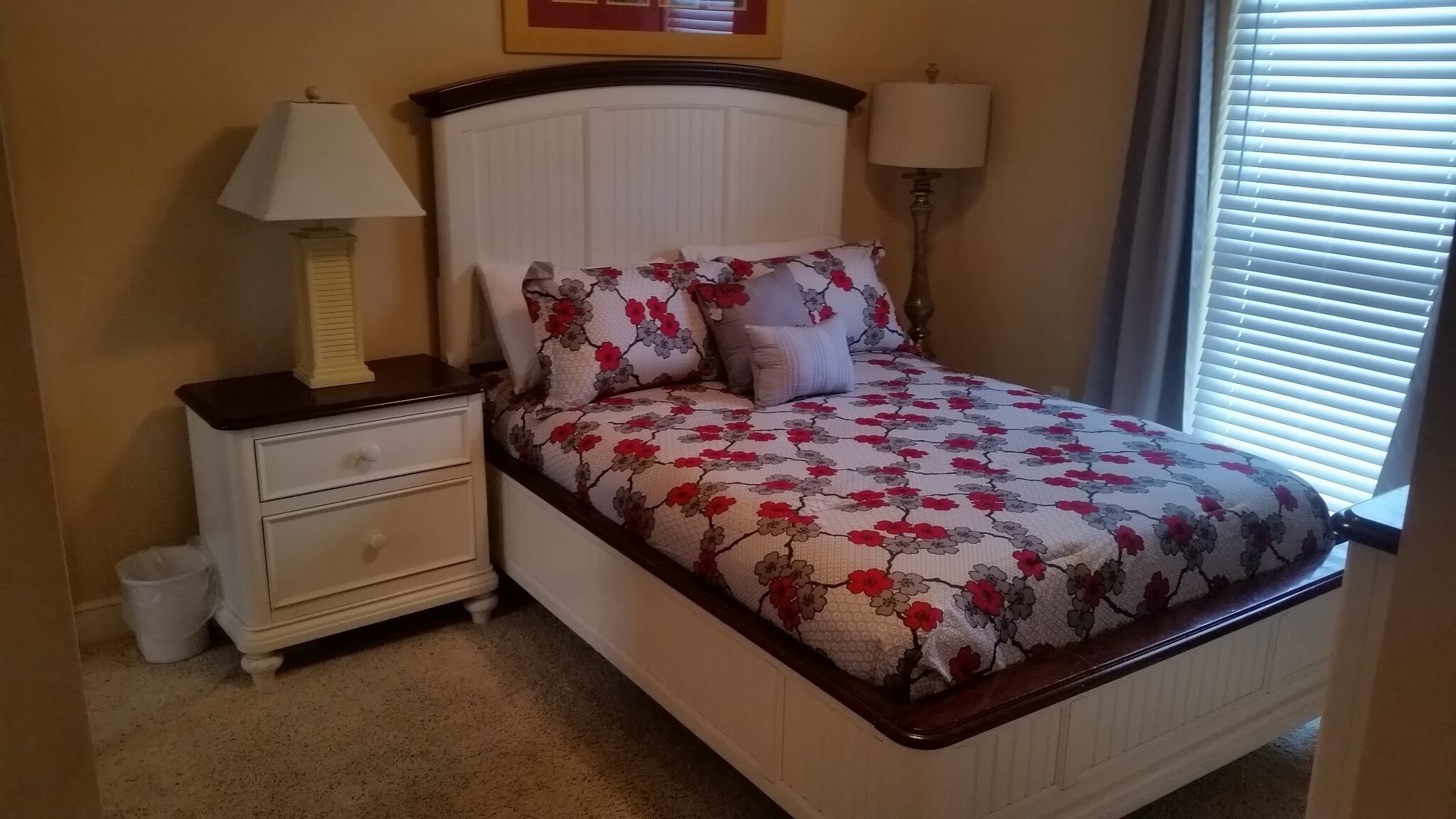 A bedroom with a bed, two bedside tables, a nightstand, a lamp, and a window with blinds.