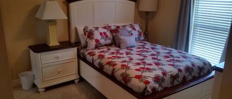 A bedroom with a bed, two bedside tables, a nightstand, a lamp, and a window with blinds.