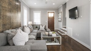 A living room with a sectional sofa, a coffee table, and a flat-screen TV mounted on the wall. - Downtown in 7min & Rooftop Deck! Gorgeous Stunning Nashville Home!  (Nashville)
