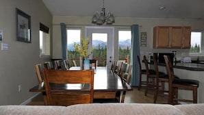 A dining area with a wooden table and chairs, a chandelier, and a view of mountains through large windows. - 5 Star Denali Park Spacious Family Home (Healy)
