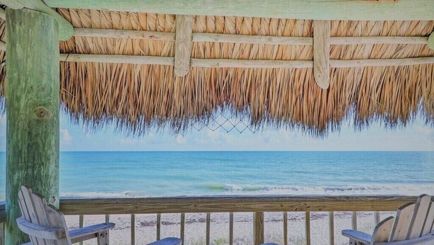 A wooden deck with two Adirondack chairs facing a beach with a thatched roof overhead.