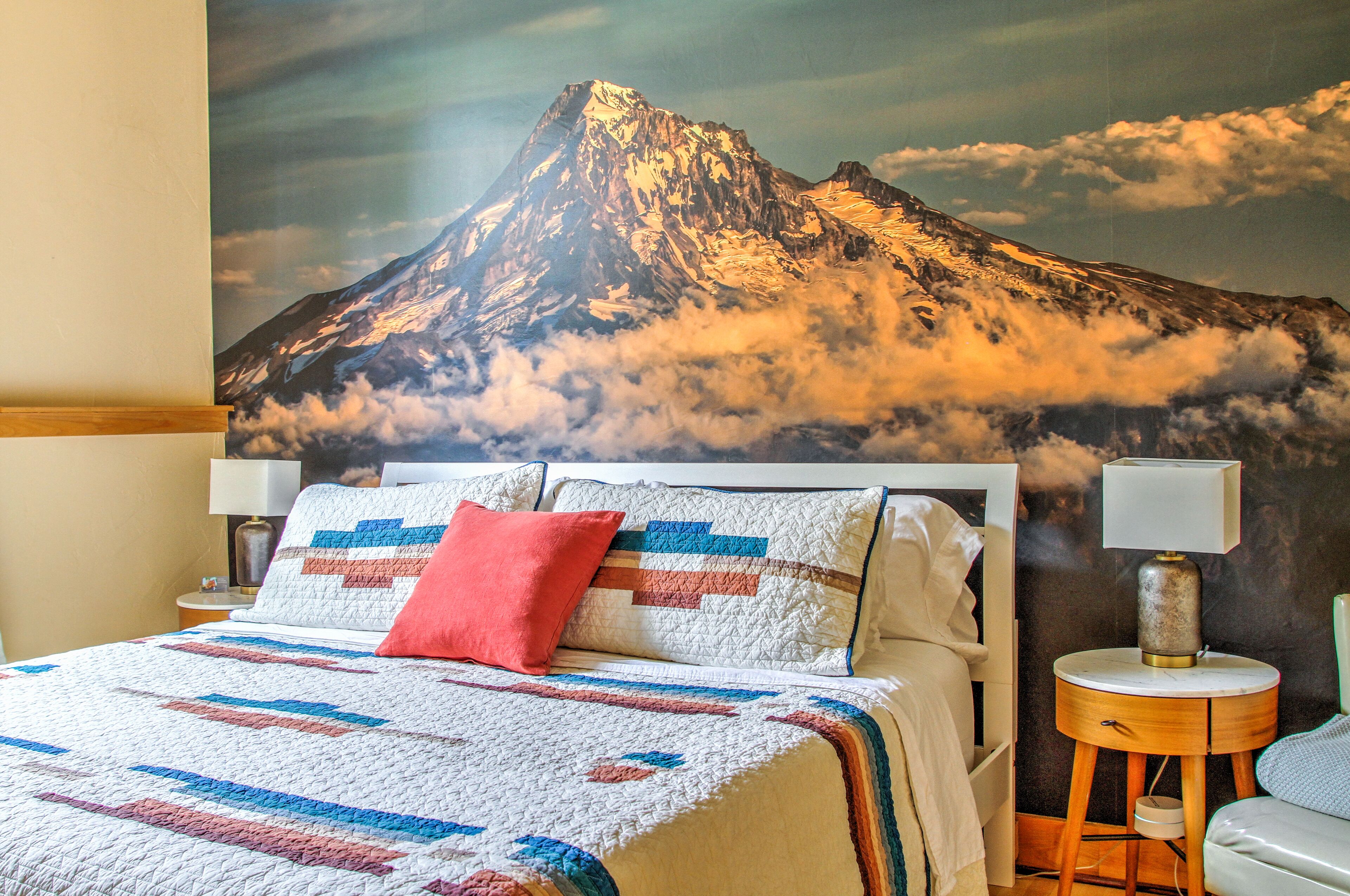 Quarto com cama, almofadas, mesa de cabeceira com abajur e um mural de parede com paisagem de montanha.