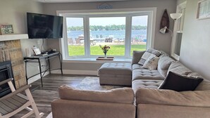 Een woonkamer met een groot raam dat uitkijkt op een meer, een flatscreen televisie, een open haard en een gezellige zithoek.