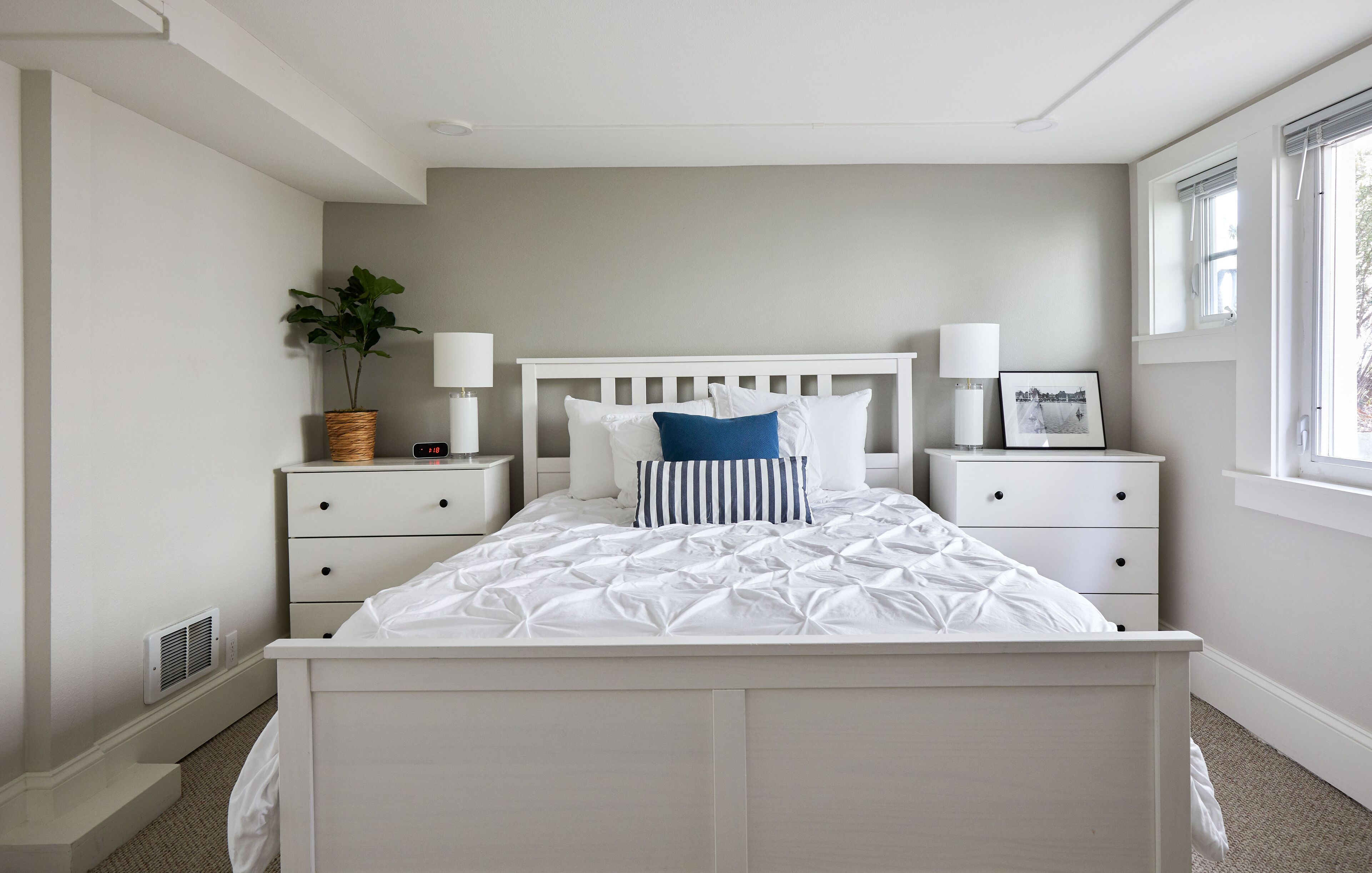 Une chambre avec un lit blanc, deux tables de chevet, une commode, un radiateur fixé au mur, une fenêtre et une plante.