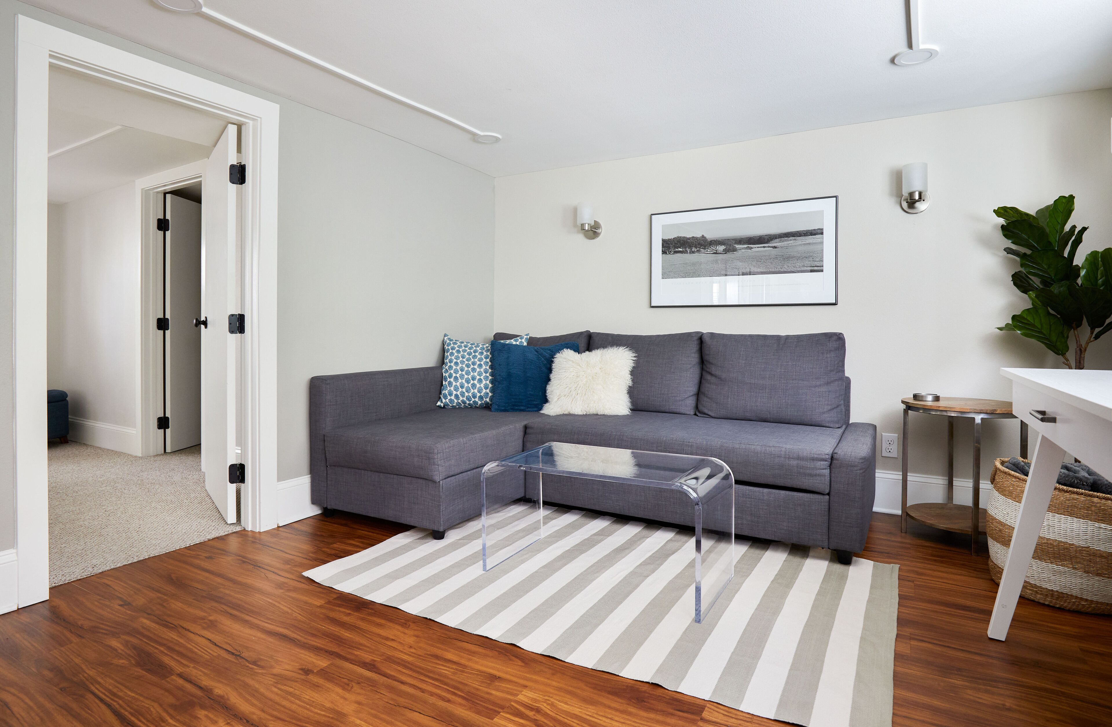 A modern living room with a grey sectional sofa, a clear glass coffee table, a striped rug, and a framed picture on the wall.